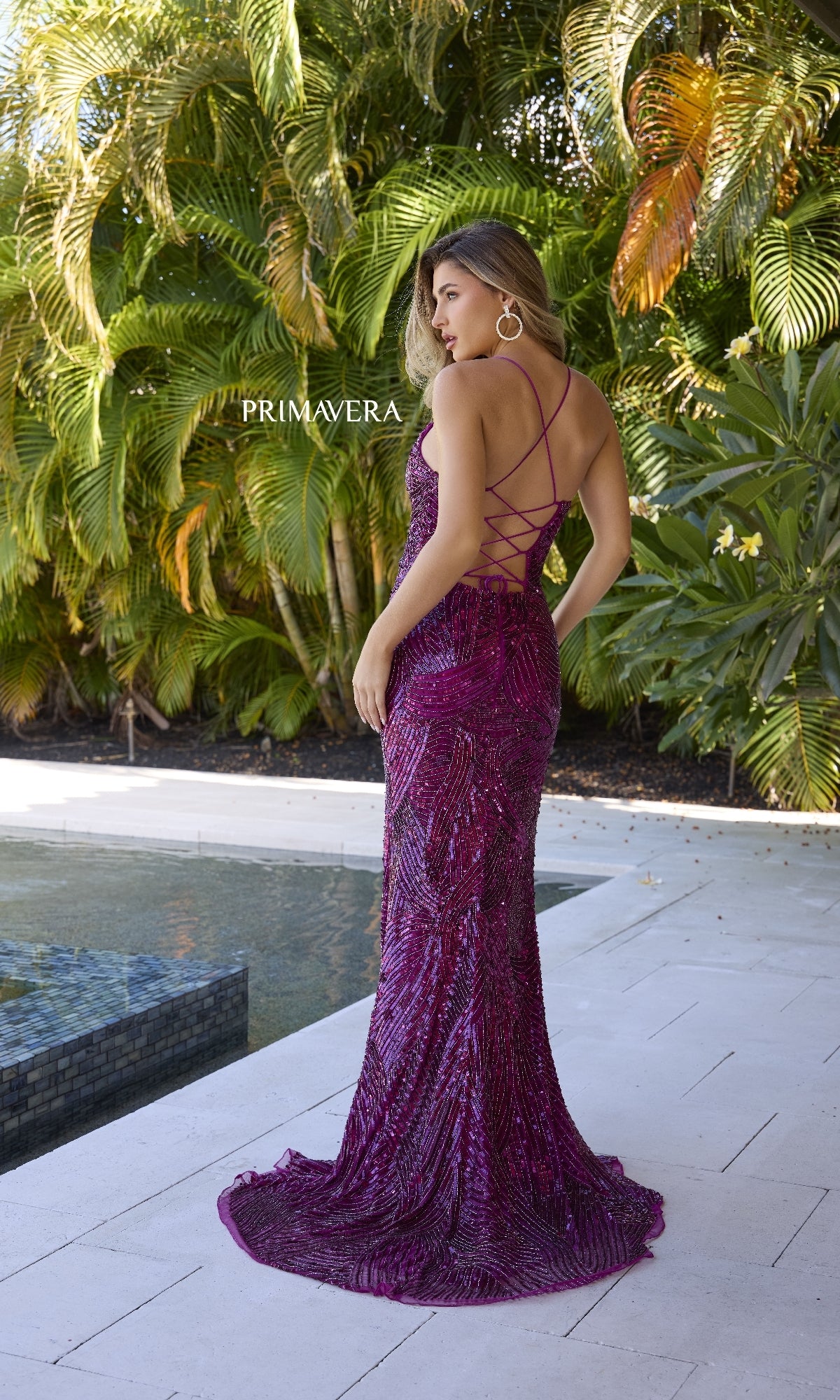 A woman stands by a pool in the Strappy-Back Long Sequin Prom Gown, Primavera 4365, featuring magenta sequins and crisscross back straps, surrounded by tropical plants.